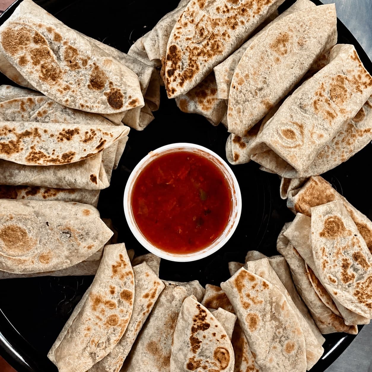 Folded burritos arranged around salsa on a black catering tray
