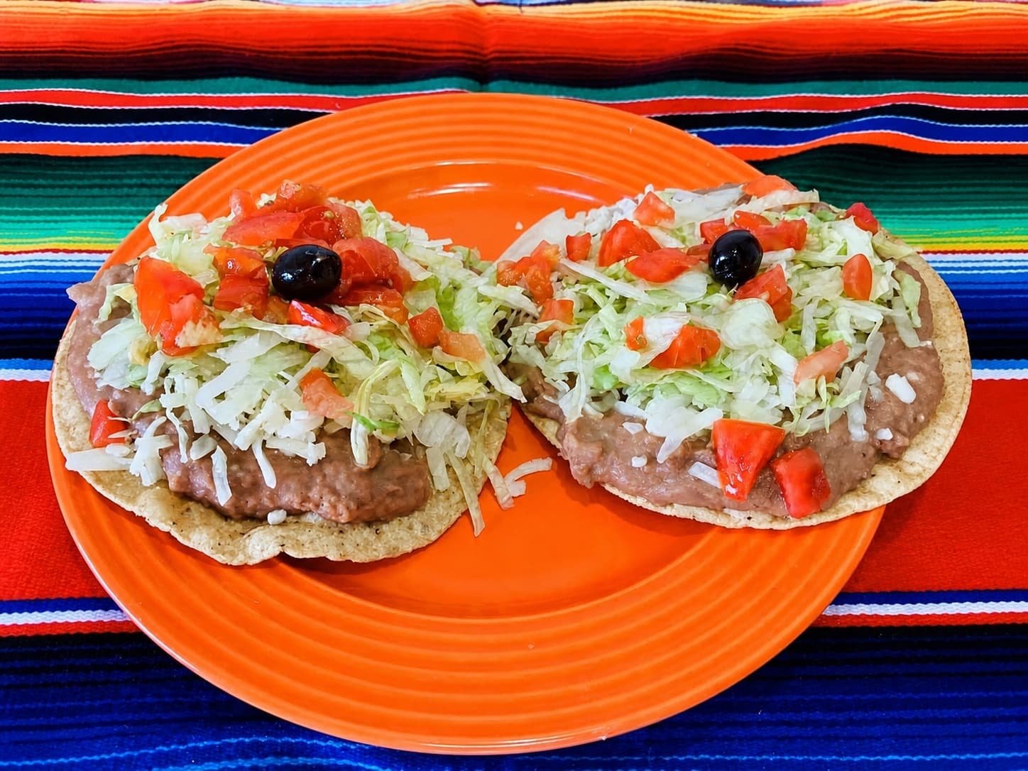 Two bean tostadas with lettuce, tomato, cheese, and olives on an orange plate